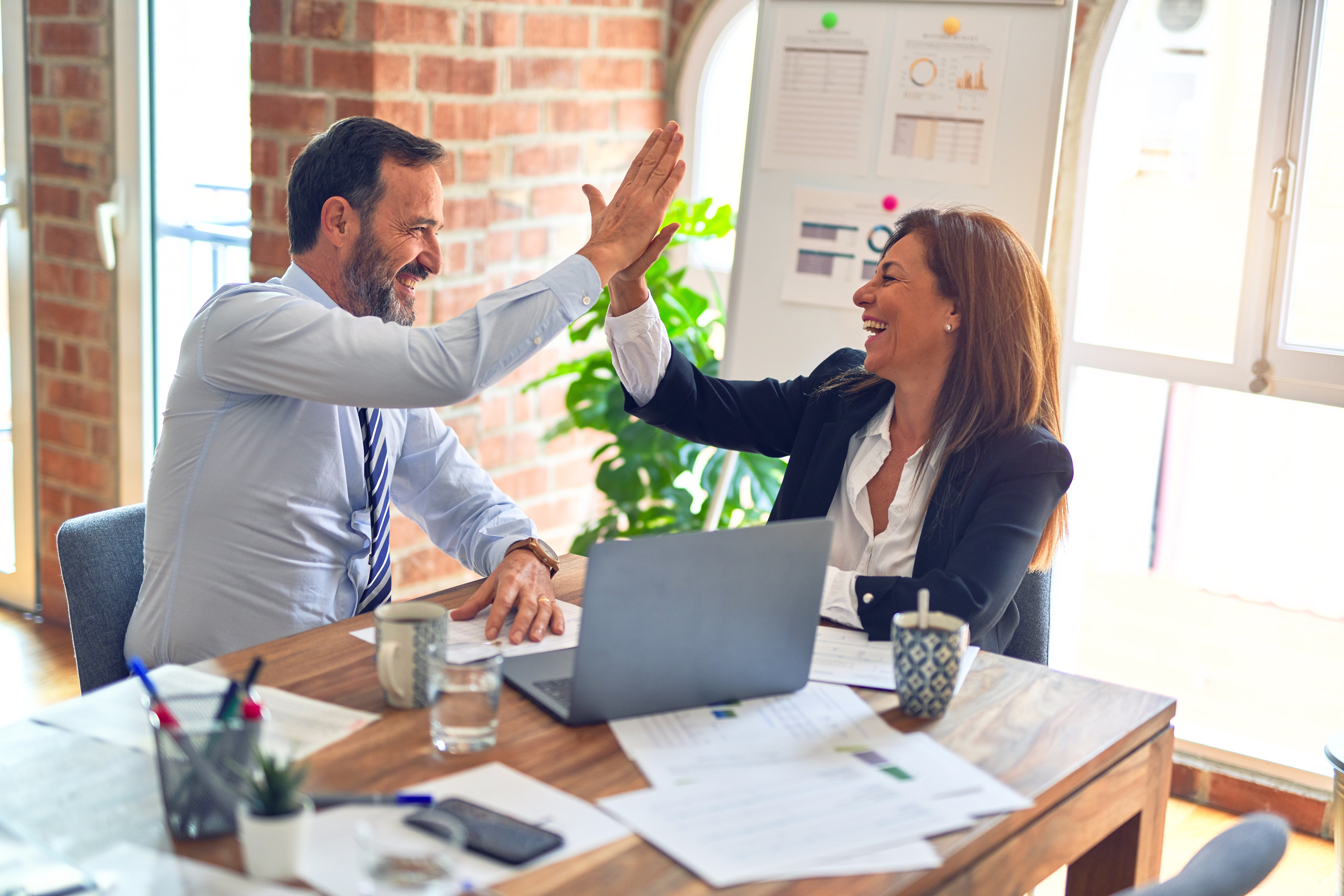 A man and woman in business attire sit at a table with a laptop in between them. They are giving each other a high five.