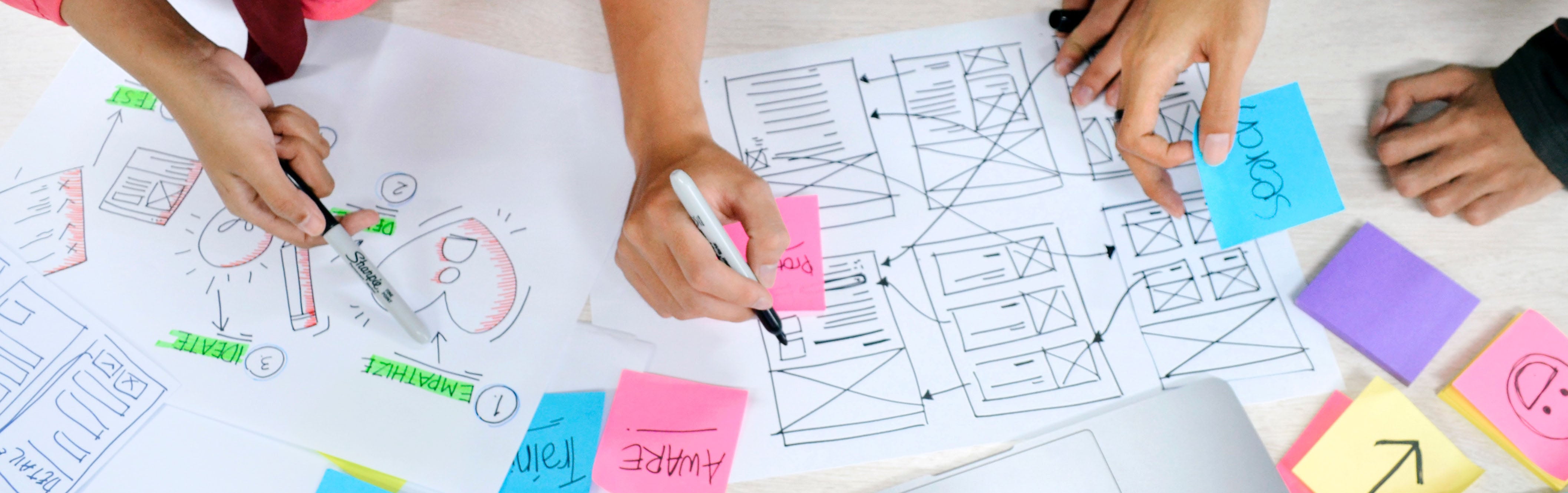 Three pairs of hands are shown resting on a table covered in papers. One hand is drawing an image with a marker. Another hand places a sticky note reading on the papers. A third hand points with a marker.