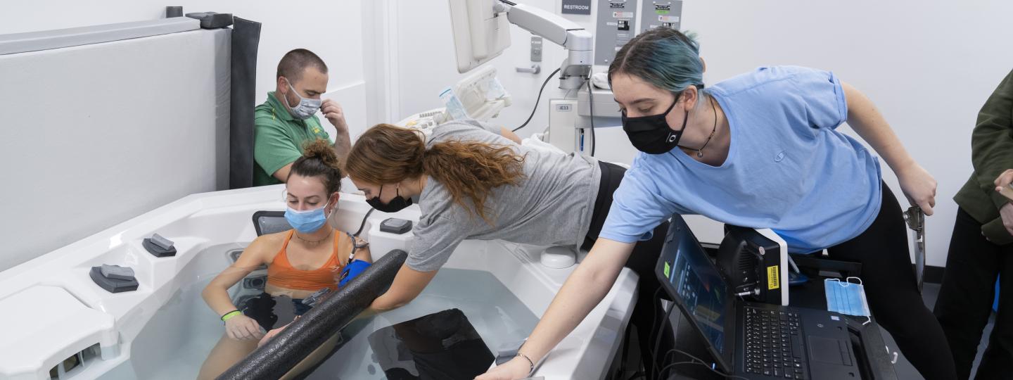 A woman sits in a shallow pool while a researcher takes her blood pressure, a second researcher adjusts the pool settings, and a third researcher enters data.