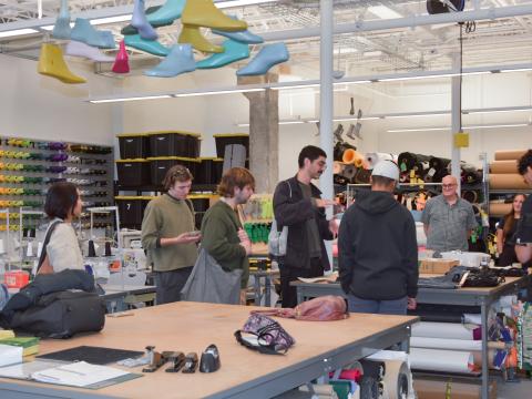 Students gather around a table in a product design lab. Rows of colored thread, shoe molds, and rolls of material are visible in the background.