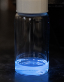 A small, clear bottle with a white cap and a small amount of glowing blue liquid in the bottom against a black background.