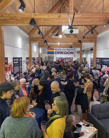 A crowded room at the opening night of the Outliers and Outlaws exhibit. A banner in the background reads "Victory over bigotry Oregon"