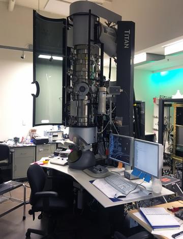 A transmission electron microscope sits on a table in a lab next to a computer.