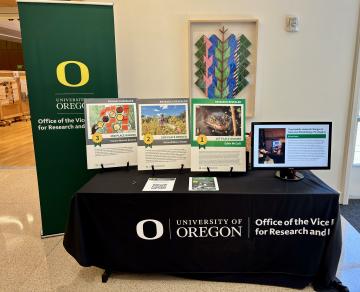 A table covered in a tablecloth that reads Office of the Vice President for Research and Innovation atop which sit three posters with images on them of a frog, a woman standing in a field of wildflowers, and a person’s hand adjusting images that are taped to a window.